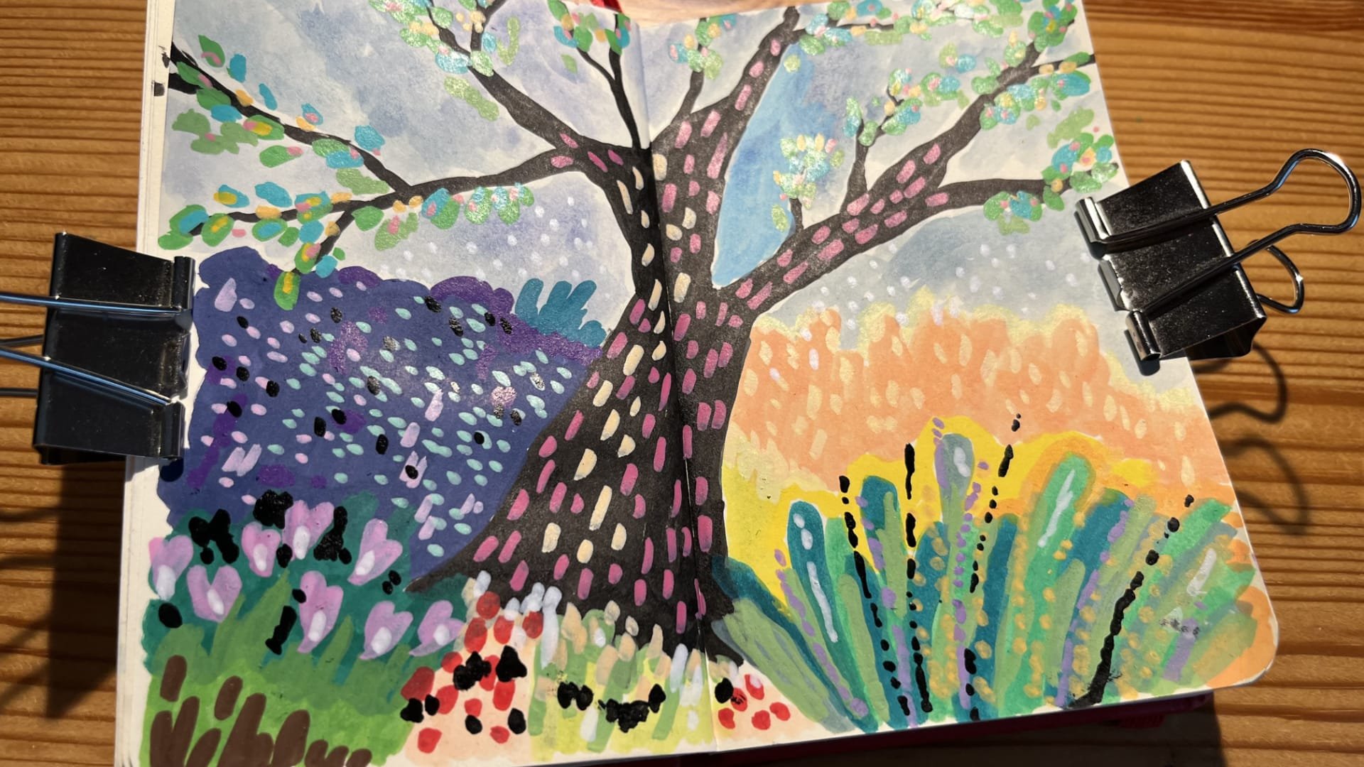Photo of an open sketchbook on a wooden table with the sketchbook spread filled with a colourful acrylic marker and watercolour drawing of a magical looking tree with black trunk and branches with pink and light pink dots on the trunk and green and pink and yellow leaves and dots on the branches, surrounded by colourful flowers and plants and with a blue sky on the background.