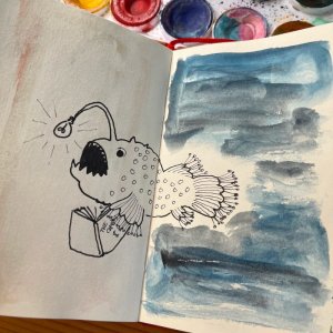 A photo of an open sketchbook spread with a drawing of a whimsical lamp fish with a lightbulb in the place of the usual “lamp” mouth open holding a book with “THE CAPITAL” and a dollar sign on the cover. On te first page, in the direction the fish is swimming, there’s a bit of metallic golden watercolour and on the second page dark blue and black watercolour by Mervi Eskelinen.