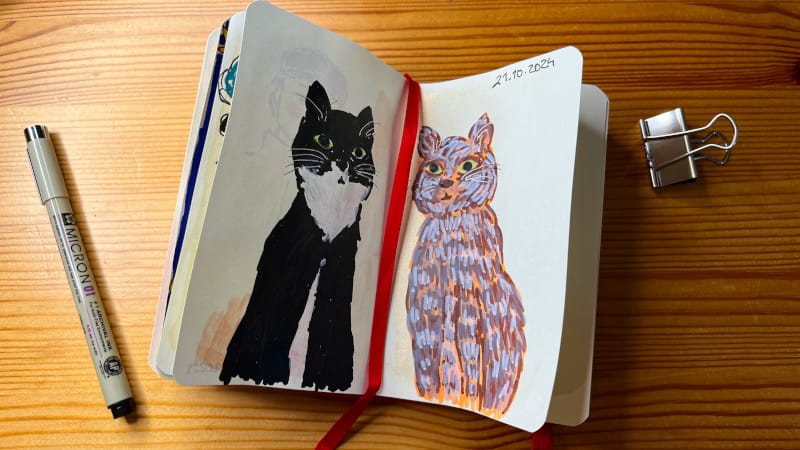 A photo of an open sketchbook on a wooden table with an acrylic marker drawing of a black tuxedo cat and brown gray tabby cat sitting looking straight at you (there's also a Sakura Micron fine liner on the table as well as a metallic clip) by Mervi Eskelinen